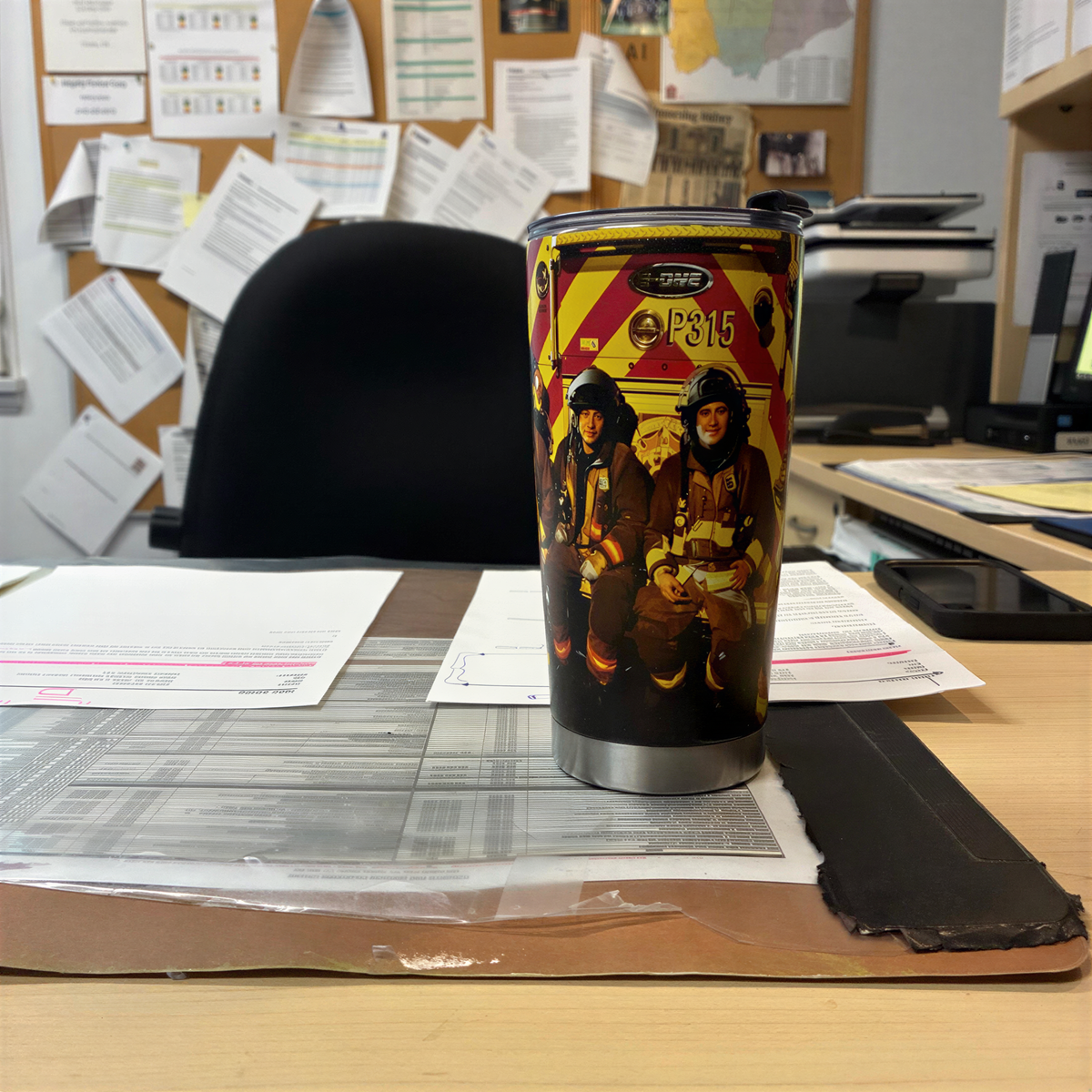 A firefighting-themed tumbler cup sits on a cluttered desk, surrounded by various papers and office supplies.